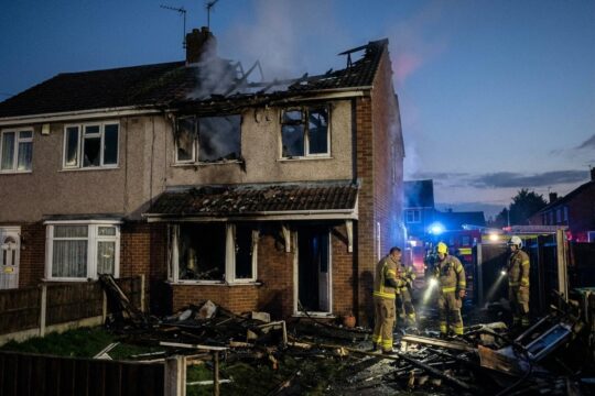 Zerstörte Doppelhaushälfte nach Hausbrand in Schermen. Dach eingestürzt, Fenster verkohlt. Feuerwehrleute inspizieren Trümmer bei Blaulicht.