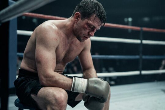 Ein nachdenklicher, erschöpfter Boxer sitzt in seiner Ringecke, der Kopf gesenkt. Sein Boxhandschuh ruht auf dem Knie, das Gesicht zeigt die physische Belastung der Nick Ball Niederlage Analyse.