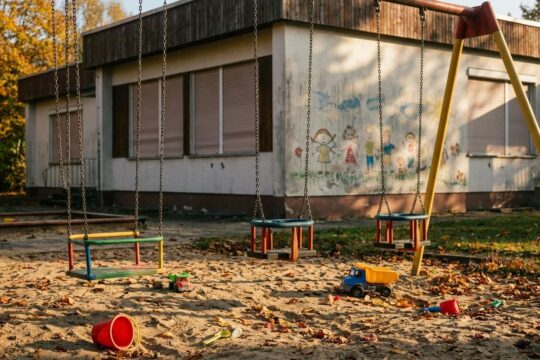 Leerer Spielplatz einer Kita mit leicht schaukelnden Schaukeln, Herbstlaub und verstreuten Spielsachen, nach der Kita Schließung Gera.