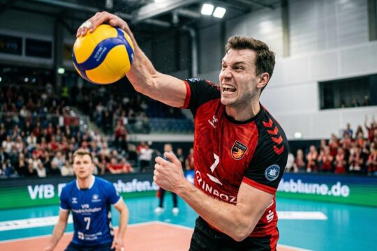 Ein BR Volleys Spieler schmettert den Volleyball hoch in der Luft, sein Blick fest auf den BR Volleys Sieg Friedrichshafen gerichtet. Gegner im Hintergrund resignieren.