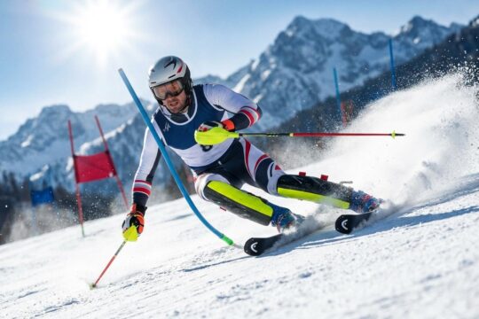 Dynamischer männlicher Ski Alpin Rennläufer in Team-Kombinations-Ausrüstung carvt mit Schneeexplosion. Präzise Technik und Geschwindigkeit prägen die Szene unter Sonne und Bergen.