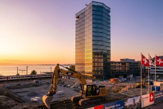Aktive Baustelle in Rorschach mit Bagger und gläsernem Hochhaus, dessen Fassade den Sonnenuntergang am Bodensee spiegelt. Der neue Stadtteil Rorschach wächst.