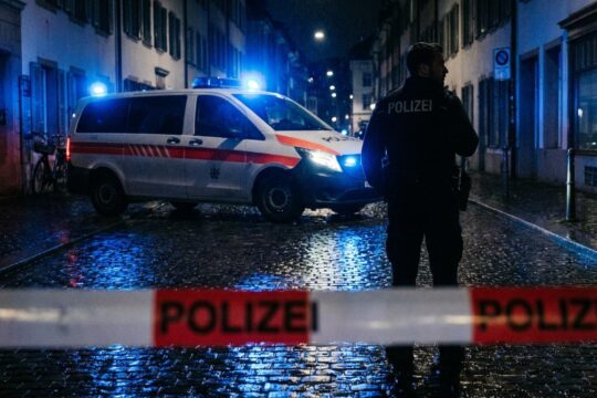 Nasse Strassen spiegeln blaue Polizeilichter in einer nächtlichen Stadt. Ein Polizist hinter Absperrband, passend für Polizeimeldungen Region Basel.