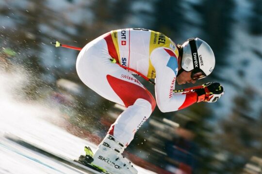 Ein Skirennläufer in extrem aerodynamischem weißem Skianzug mit roten Akzenten rast den Hang hinab. Die Farbe und hohe Geschwindigkeit sind deutlich sichtbar.