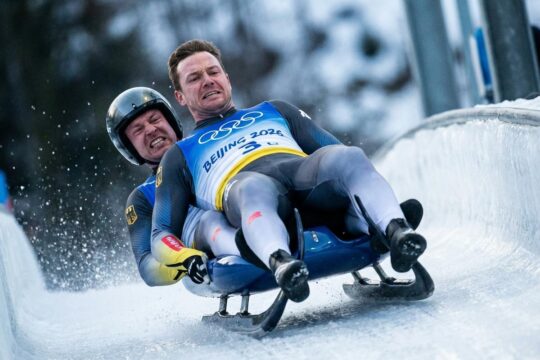 Zwei Rodler rasen bei Olympia 2026 Rodeln Doppelsitzer konzentriert durch eine eisige Kurve, winzige Eispartikel fliegen. Dramatische Szene.