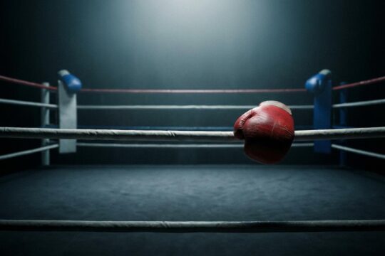 Roter Boxhandschuh auf Ringseil, blauer Handschuh entfernt im Schatten. Die leere, spärlich beleuchtete Ringmitte zeigt: Der Joshua Fury Kampf unsicher.