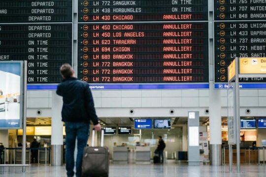 Anzeigetafel im modernen Flughafen-Terminal zeigt mehrere Lufthansa-Flüge in Rot als 'ANNULLIERT'. Ein Reisender steht ratlos mit Koffer davor und erlebt die Auswirkungen des Lufthansa Streiks.