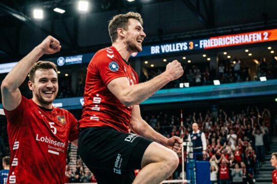 BR Volleys Spieler jubeln ausgelassen nach dem dramatischen BR Volleys Sieg Friedrichshafen. Einer springt in die Luft, der andere lächelt, Fans im Hintergrund.