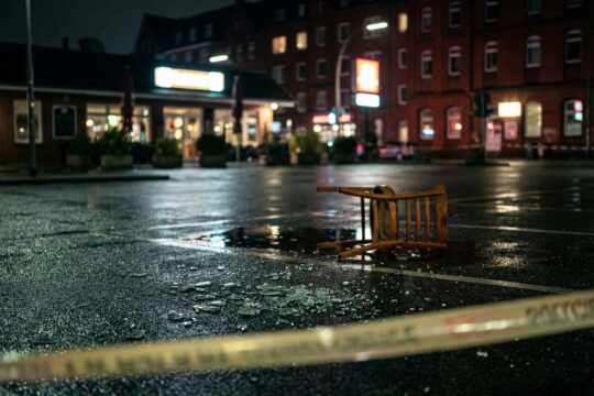Nasser Hamburger Parkplatz bei Nacht: Zerbrochenes Glas, umgestürzter Stuhl und Polizeiband symbolisieren einen tödlichen Streit in Hamburg.