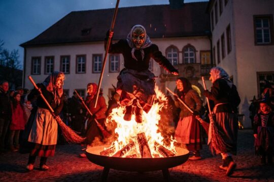 Beim Hexensprung Bad Bergzabern springt eine Oberhexe dynamisch über lodernde Flammen. Maskierte Hutschelhexen tanzen, Schloss Bad Bergzabern im Dämmerlicht.