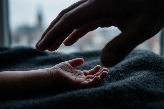 Eine winzige Babyhand liegt schutzlos, während sich bedrohlich eine große Männerhand nähert. Ein dunkler Schatten fällt auf die Hand in Halle und symbolisiert die Misshandlung des Babys.
