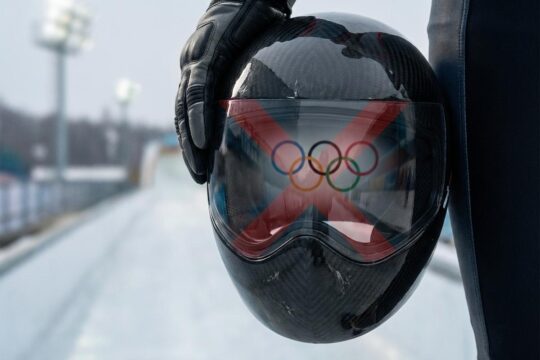 Ein dunkler Skeleton-Rennhelm, von behandschuhten Händen gehalten. Das blockierte Olympia-Logo im Visier verdeutlicht das Heraskewytsch Olympia Startverbot.
