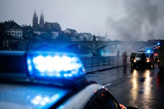 Intensive Blaulichter eines Einsatzfahrzeugs auf nassem Asphalt. Im nebligen Hintergrund die Basler Architektur – ein Sinnbild für das Blaulicht Region Basel.