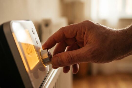 Eine Hand stellt achtsam den Drehregler eines digitalen Thermostats ein, um die Heizung richtig einzustellen und Energie zu sparen. Der warme Wohnraum im Hintergrund suggeriert Behaglichkeit.