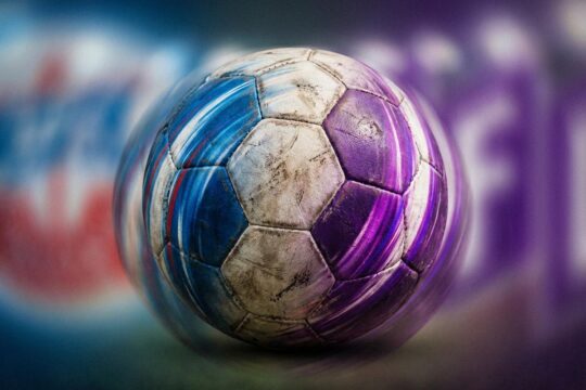 Ein abgenutzter Fußball spiegelt die Vereinsfarben von Hansa Rostock und VfL Osnabrück wider, umgeben von Stadionlicht-Drama des späten Remis.