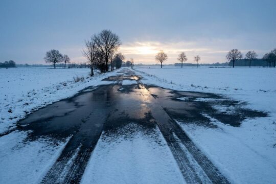 Eisglatte Landstraße in Niedersachsen bei Dämmerung. Schnee und spiegelndes Eis prägen die Fahrbahn, Reifenspuren warnen vor der Gefahr durch Schnee und Glätte.
