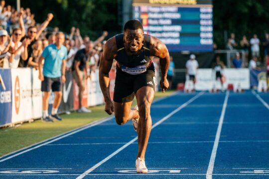 Ein sprintender Athlet mit angespannter Muskulatur überquert auf einer blauen Tartanbahn die Ziellinie. Jubelnde Zuschauer erwarten die aktuellen Leichtathletik Ergebnisse.