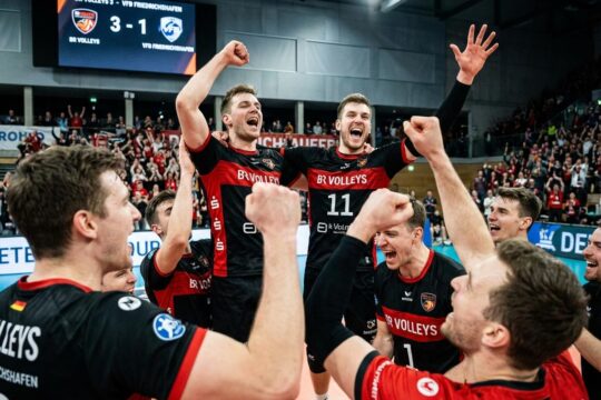 Der BR Volleys Sieg Friedrichshafen: Spieler in schwarz-roten Trikots jubeln mit erhobenen Armen und geballten Fäusten. Ihre Gesichter strahlen pure Freude und Triumph in der hell beleuchteten Sporthalle aus.