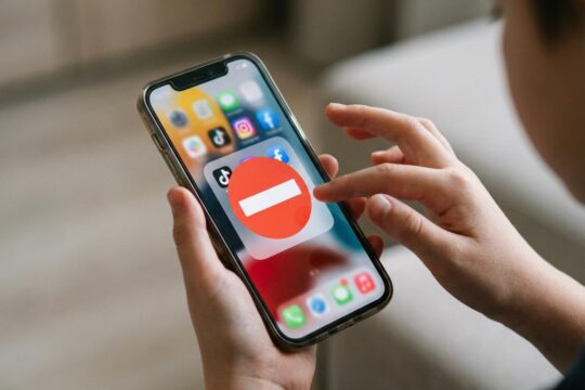 Kinderhände halten ein Smartphone, auf dessen Display ein rotes Verbotsschild Social-Media-Logos überdeckt. Dies visualisiert ein Social Media Verbot für Minderjährige.