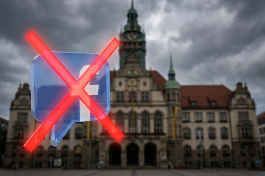 Ein prominentes rotes X über einem blauen Facebook-Kommentar-Icon vor einem unscharfen Stadtverwaltungsgebäude visualisiert, wie die Stadt Facebook Kommentare sperrt.