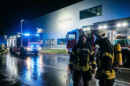 Professioneller Feuerwehreinsatz bei Wagner Pizza: Einsatzkräfte mit Atemschutz vor blau blinkenden Fahrzeugen am nassen Werk.